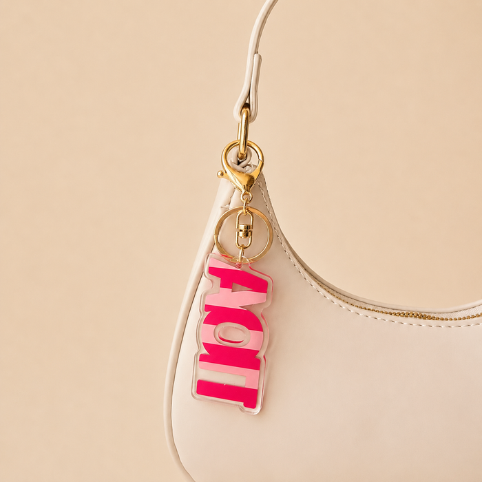 A close-up of a white handbag with a gold clasp and The Darling Effectβs Alpha Omicron Pi Stripes Acrylic Keychain attached. The pink and white layered letters spell out "LOVE." The soft beige background adds a gentle touch. Perfect sorority gift!.