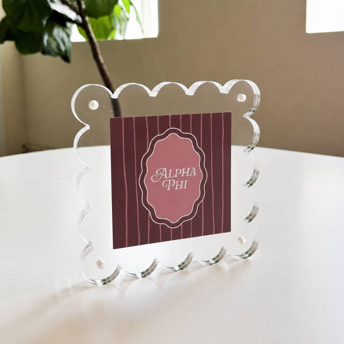 The Darling Effectโs Alpha Phi Square Scalloped Acrylic Frame, featuring a clear scalloped-edge design, displays a pink and maroon Alpha Phi card. It sits magnetized on a white table near a green plant with windows in the background.