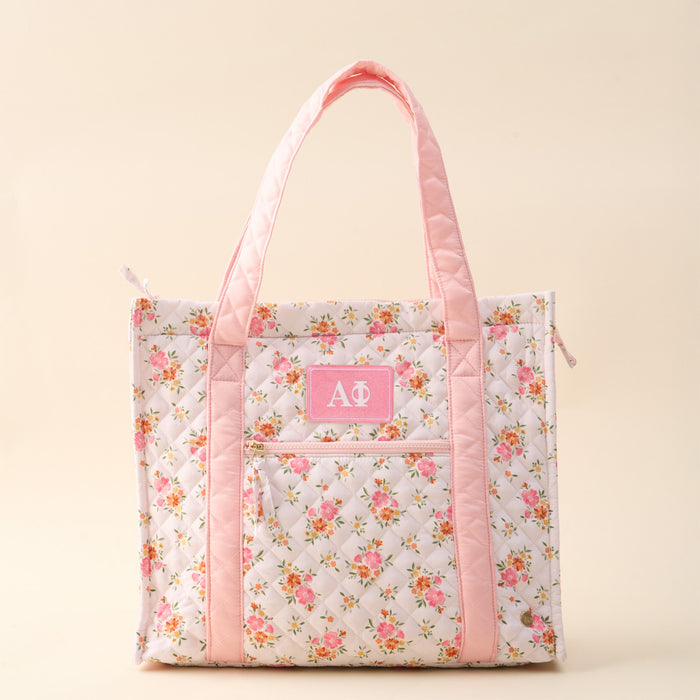 The Darling Effect's Alpha Phi The Everyday Quilted Tote Bag features a floral quilted design, light pink handles and trim, a front pocket with white Greek letters on a pink patch—ideal for displaying your sorority pride in style.