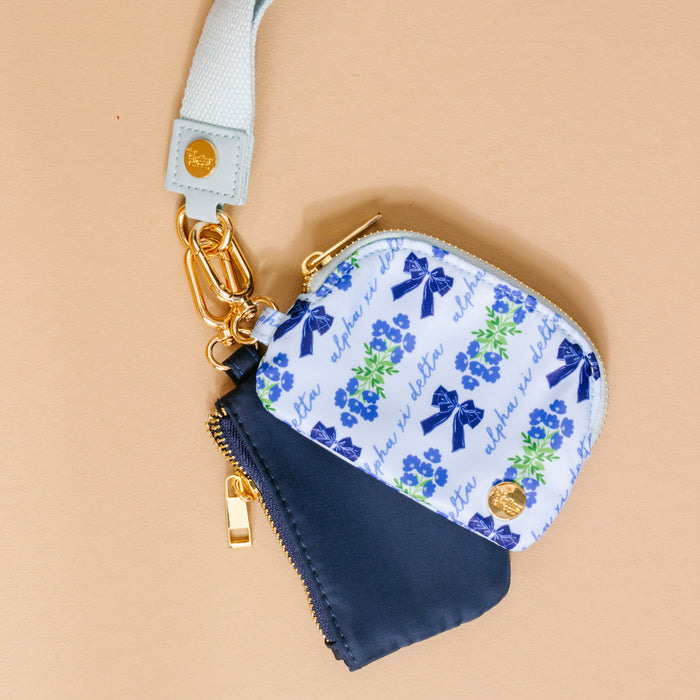 The Darling Effect’s Alpha Xi Delta Dynamic Duo Pouch Wristlet features two small zippered pouches—one navy, one white with blue bows and green flowers—on a light blue strap, set against a beige background.