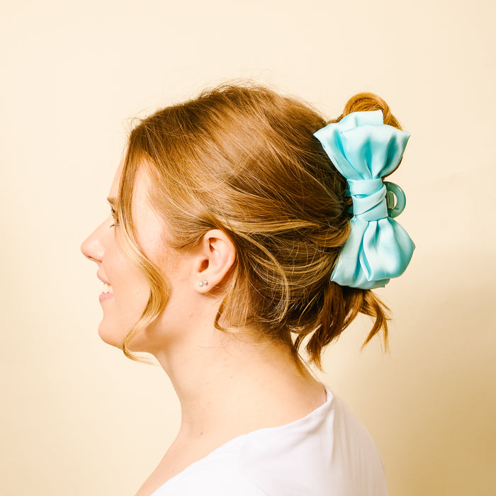 A woman with light brown hair smiles in profile, wearing a white shirt and securing her updo with the-darling-effect Aqua Satin Bow Claw Clip against a beige background.