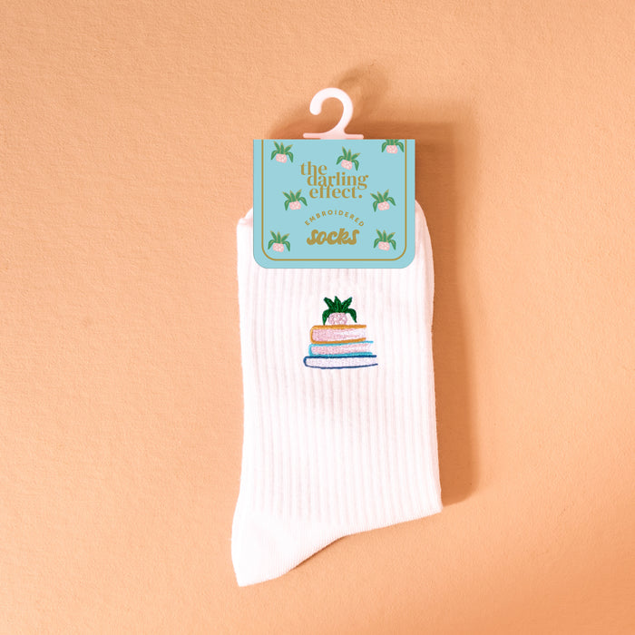 The Book Nook Embroidered Crew Socks is a white ribbed women’s sock featuring embroidered books and a small green plant, shown against a peach background with packaging labeled “the darling effect” on a light blue tag.