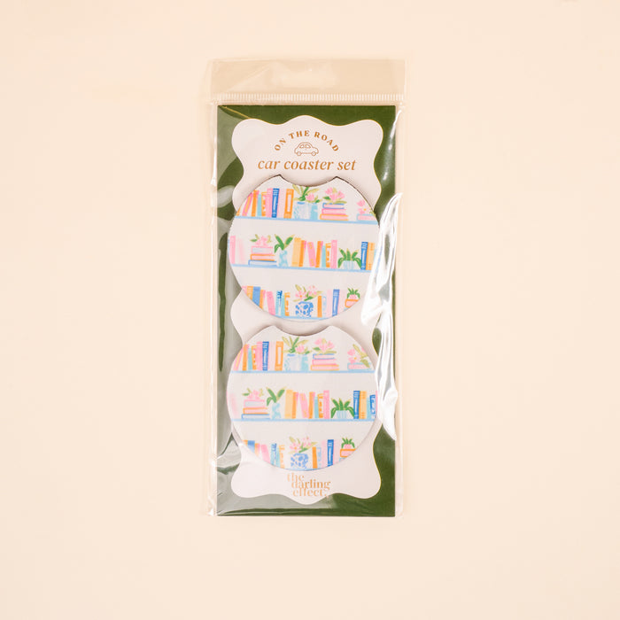 The Book Shelfie On the Road Car Coaster Set by The Darling Effect includes two absorbent coasters for your car cup holder, featuring vibrant books and plants on shelves. Packaged in clear plastic with branded labeling.