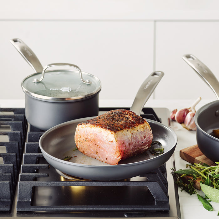 Seasoned meat cooks in a gray ceramic nonstick skillet, part of a 10-piece cookware set, with pot and herbs beside it.