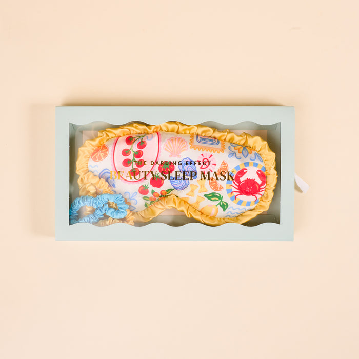 The Darling Effect Ciao Bella Beauty Sleep Mask set features a floral satin mask with yellow trim, blue satin scrunchie, and comes in a light green box with clear window. "Beauty Sleep Mask" is printed on the mask.