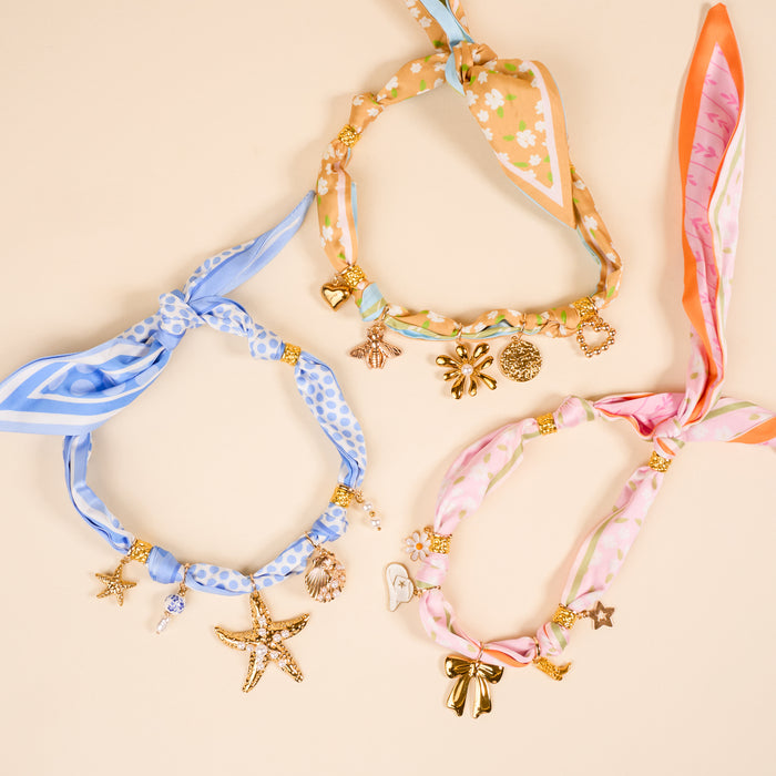 Three patterned silk scarvesβblue polka dot, orange floral, and pink flowerβare adorned with The Darling Effect's Coquette Cloud The Ultimate Charm Pack: gold stars, hearts, bows, and shells on a beige background.