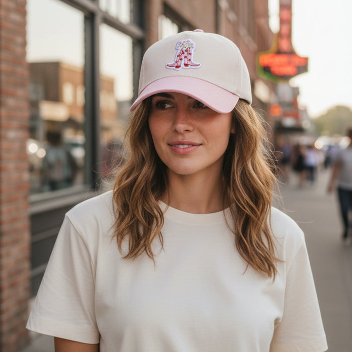 A woman wears a cream t-shirt and The Darling Effect Cowgirl Boots Classic Canvas Hat, featuring a relaxed fit, tan canvas, pink brim, and pink cowboy boots patchโperfect for everyday wear.