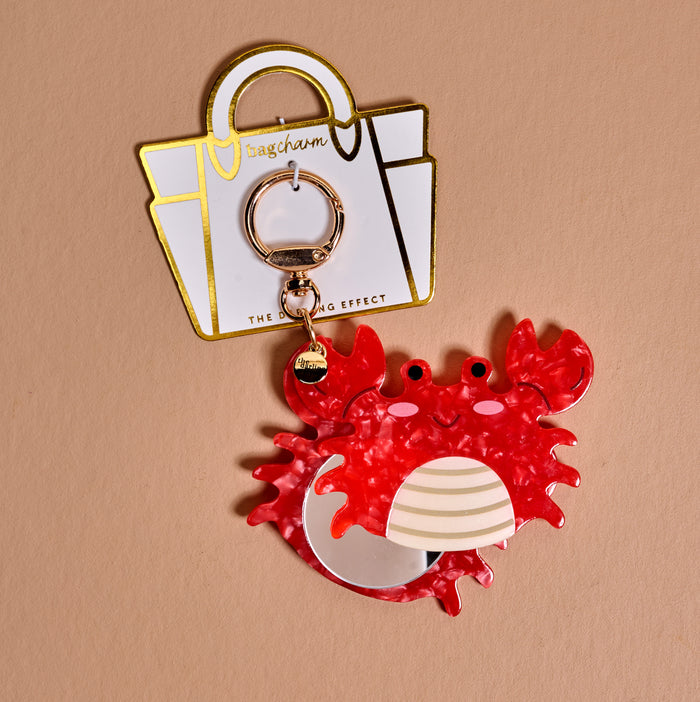The Darling Effect Crab Mini Mirror Bag Charm features a red crab-shaped keychain with an acrylic mirror, attached to a gold keyring on a white and gold cardβa cute accessory for your bag, shown on a light brown background.
