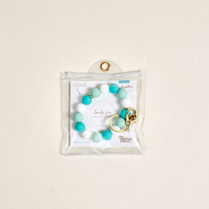 The Darling Effectβs Daydreamer Hands-Free Keychain Wristlet features teal and white silicone beads with a gold keyring, packaged in a clear pouch against a light beige backgroundβstylish convenience for on-the-go use.