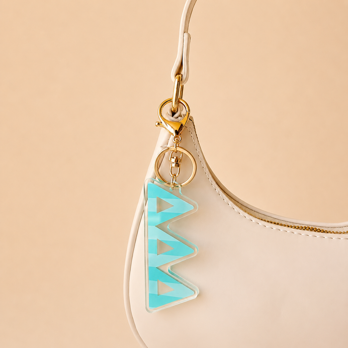 A cream-colored handbag with a gold clasp features The Darling Effectβs Delta Delta Delta Stripes Acrylic Keychain in turquoise and clear zigzags on the handleβperfect as a stylish bid day gift. The background is plain light beige.