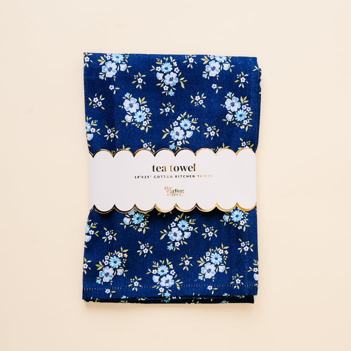 The Endless Daydream Navy Tea Towel by the-darling-effect features small blue and white flowers, is folded with a scalloped โtea towel, 18x28โ cotton kitchen towel label, and set against a light beige background.