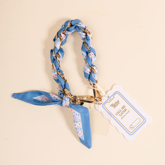 The Darling Effectβs Forget Me Not Call Me Later Phone Scarf features a blue floral braid with a gold chain, looped in a knot with secure grip and gold clasp, plus a white and gold Call Me Classy tag, displayed on a light beige surface.