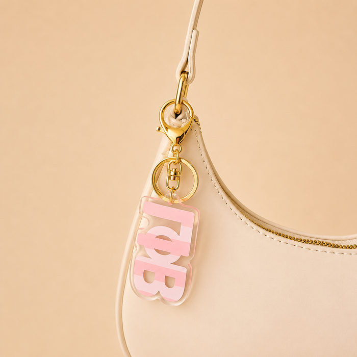 A cream-colored handbag with a gold zipper features The Darling Effect's Gamma Phi Beta Stripes Acrylic Keychain in pink and whiteβan ideal sorority gift.
