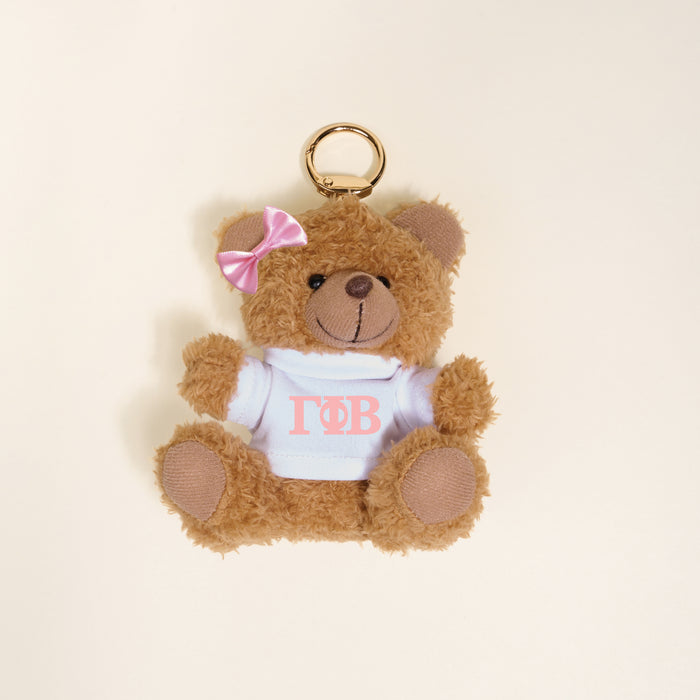 A small brown Gamma Phi Beta Teddy Bear Bag Charm by The Darling Effect, with a pink bow and white shirt with pink Greek letters, shown on a light beige backgroundβideal as a cute sorority accessory or bag charm.