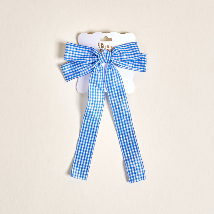 The Darling Effect Gingham Blue Double Take Hair Bow features blue and white gingham with long tails, perfect for hair accessories, attached to a scalloped-edge white display card on a light beige background.