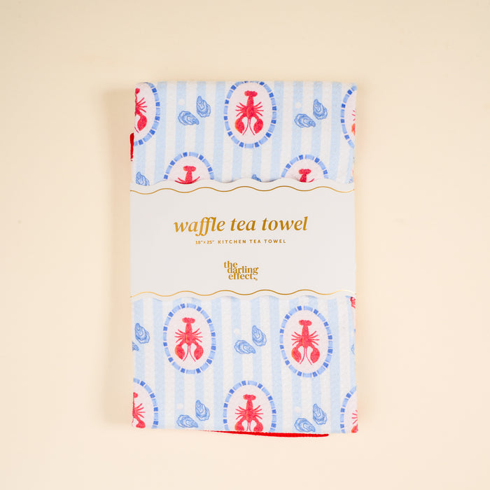 A folded Harbor House Waffle Tea Towel by The Darling Effect features a blue and red lobster and seashell pattern, wrapped in a label, displayed on a light beige background.