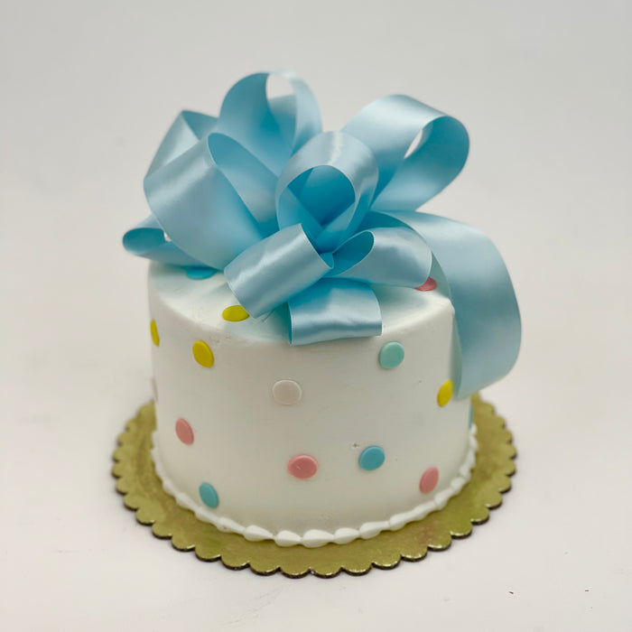 A decorated cake with white fondant, pastel blue polka dots, and a blue satin bow on top, presented on a gold stand.