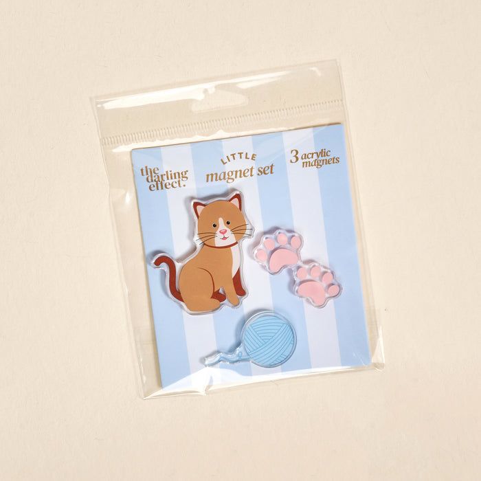 The Darling Effectβs Kitty Corner Little Magnet Set features acrylic magnets of a brown cat, pink paw prints, and a blue yarn ball on a blue and white striped backgroundβperfect for decorating your fridge.