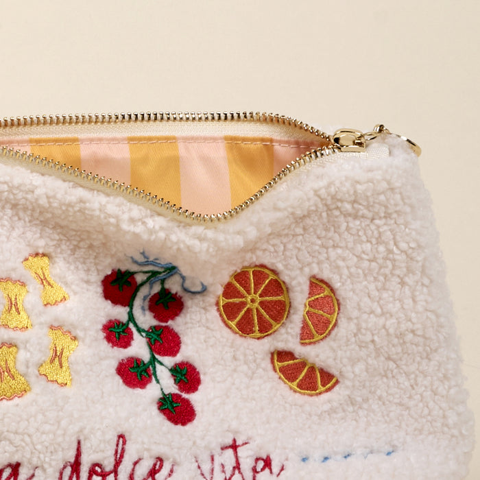 A close-up of The Darling Effectโs La Dolce Vita Teddy Pouch shows fuzzy white sherpa fabric embroidered with tomatoes, oranges, and pasta, and a partially open zipper revealing its pink and yellow striped lining.