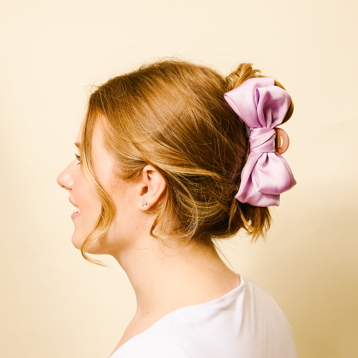 A woman with light brown hair in a loose bun, secured with the-darling-effect's Lilac Satin Bow Claw Clip, smiles and faces left against a light beige background. She wears a white top, highlighting this chic accessory for easy updos.