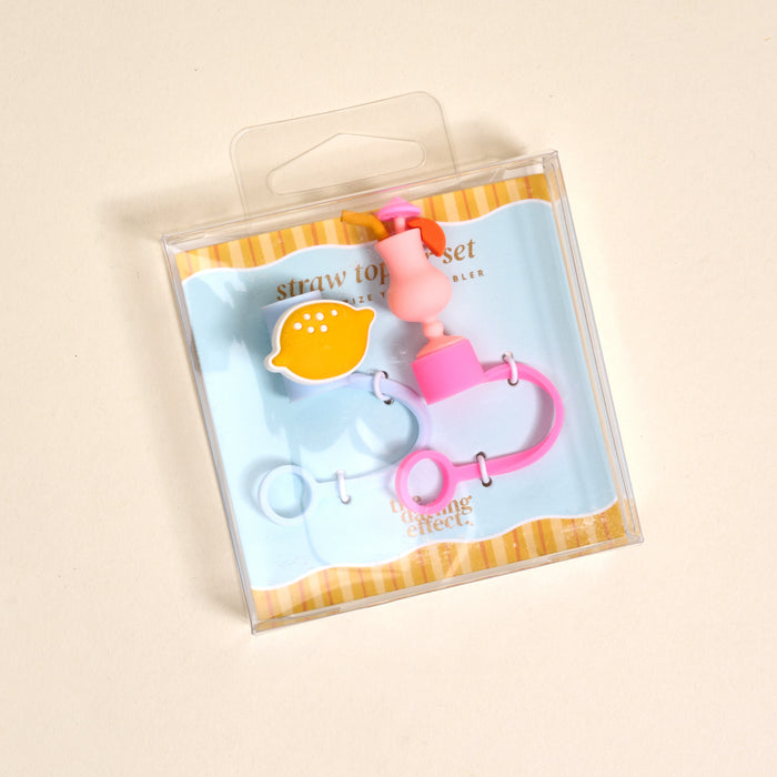 The Darling Effectβs Limoncello Love Straw Topper Set includes leak-resistant pink and yellow lemon-shaped and pink ring silicone toppers, adding fun to your drinkware accessories. Displayed on a light blue and yellow striped background.