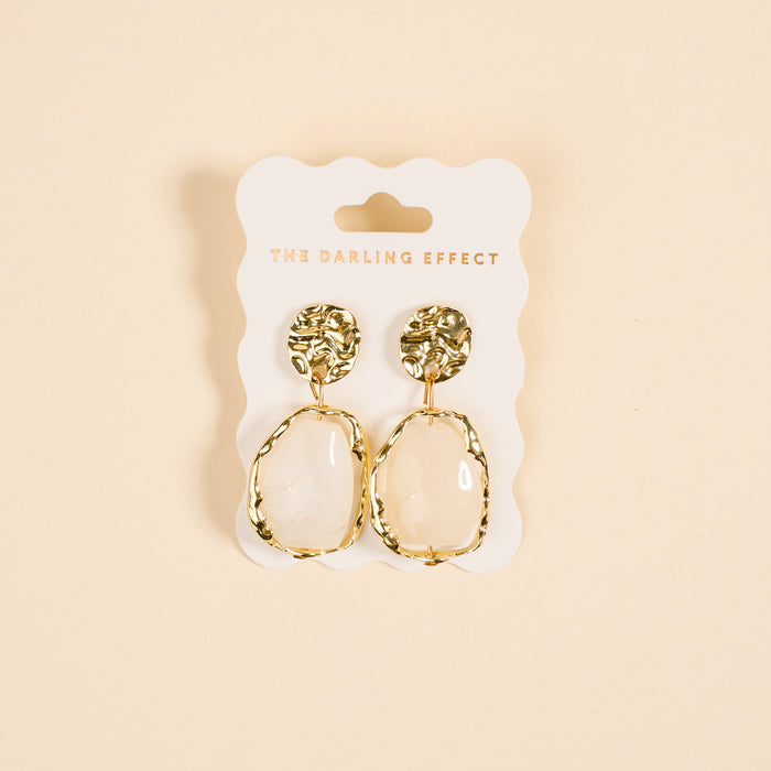 The Darling Effect's Liquid Gold Standout Earrings feature textured gold tops, irregular oval-shaped white stones, and a high-shine finish. Displayed on a branded card.