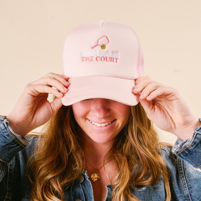 A person smiles, holding the brim of the-darling-effectβs Meet Me at the Court Trucker Hatβpink with "meet me at THE COURT," a mini tennis racket and ballβstylishly paired with a denim jacket.