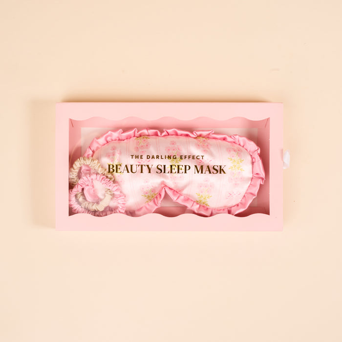 A wavy-edged pink box holds The Darling Effectβs Oh So Fancy Pink Beauty Sleep Maskβa light pink satin mask with a floral pattern and gold text: THE DARLING EFFECT and BEAUTY SLEEP MASK.