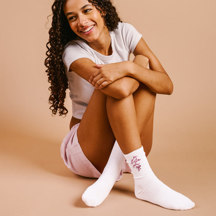 A young woman with curly hair, in a white t-shirt, pink striped shorts, and The Darling Effect's Pi Beta Phi Embroidered Crew Socks featuring red text, sits smiling on the floor against a light beige background.