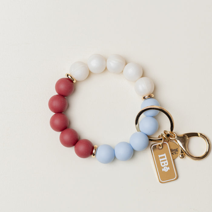The Darling Effectβs Pi Beta Phi Hands-Free Keychain Wristlet features red, white, and light blue beads with gold accents and a white logo charmβperfect as a stylish sorority keychain accessory.