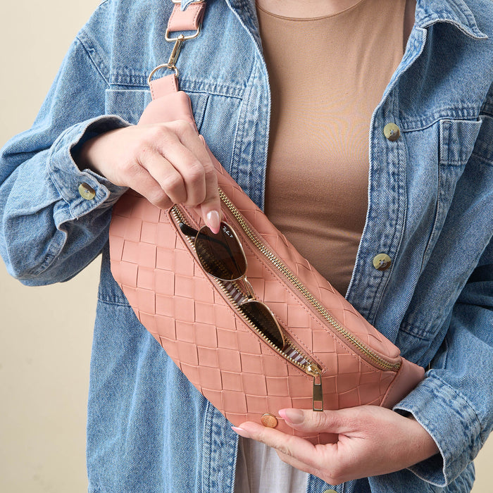 A person in a tan shirt and blue denim jacket puts sunglasses into The Darling Effectβs Rose Trendy Luxe Belt Bag, a pink woven vegan leather crossbody with a gold zipper, highlighting this fashionable accessory.