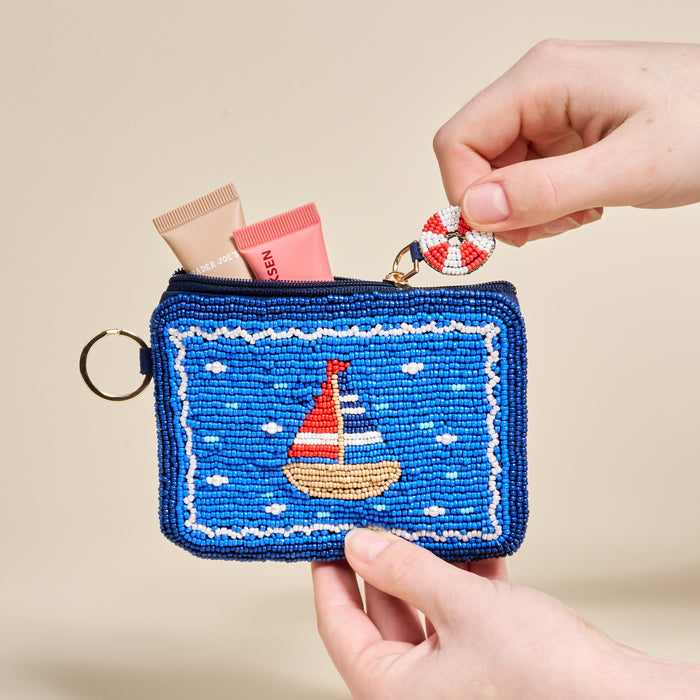 The Darling Effect's Sail Away Handcrafted Beaded Pouch in blue, held by two hands, features a sailboat design and comes with a small life preserver keychain attached to the zipper.