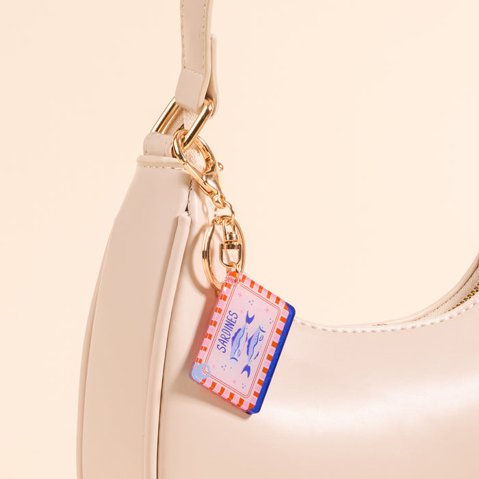 Close-up of The Darling Effect’s Sardines Acrylic Keychain, featuring pink and red stripes and fish illustrations, attached to a cream handbag with a gold clasp. The beige background enhances this stylish accessory's charm.
