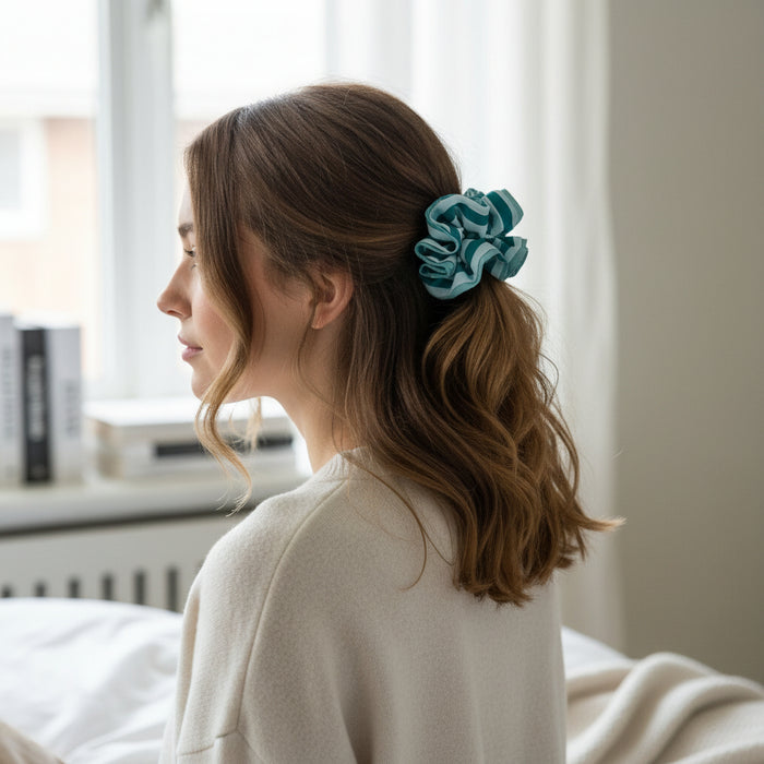A woman with wavy brown hair sits on a bed, facing left. She wears a cream top and her hair is styled in a loose ponytail with The Darling Effectโs Sicily Stripes Teal Ruffle Up Scrunchie. Soft natural light streams through the window behind her.