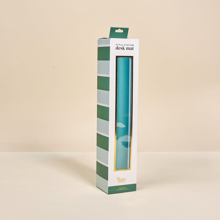The Sicily Stripes Teal Style Station Desk Mat by The Darling Effect comes in upright white packaging with green stripes and a window displaying the blue mat inside. Labeled "desk mat" at the top, it's perfect for workspace transformation.