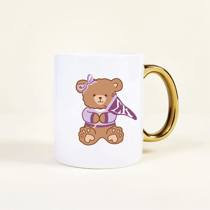 The Sigma Kappa 11 oz Cozy Cute Ceramic Mug by The Darling Effect features a brown teddy bear in a purple bow and sweater, holding an โLKโ pennant. The white mug has a gold handle for stylish flair.
