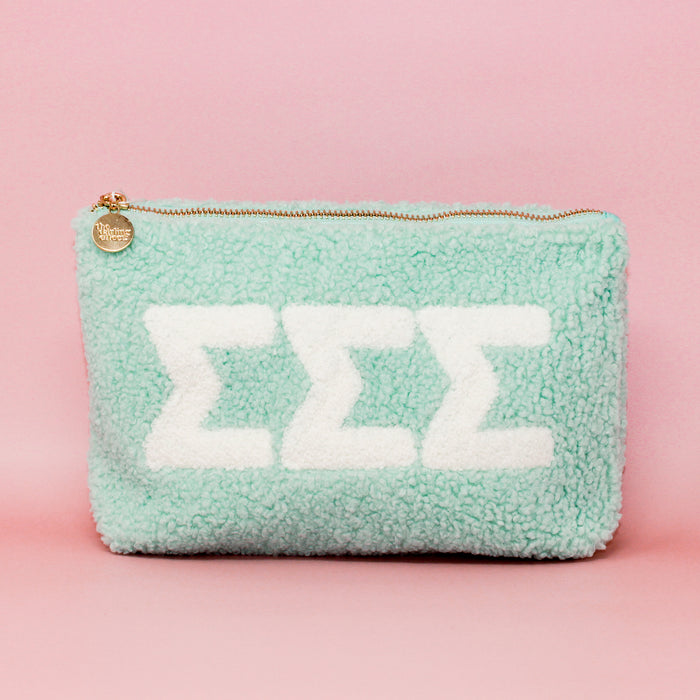The Darling Effectβs Sigma Sigma Sigma Aqua Teddy Letter Pouch features plush mint sherpa, a gold zipper, round tag, and three white ΣΣΣ lettersβideal as a chic carry-on accessory. Styled with a light pink background.
