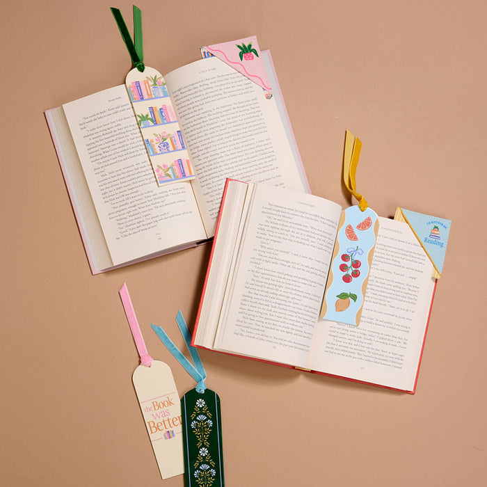 Three open books are arranged on a beige surface, each marked with The Book Was Better Acrylic Bookmark by The Darling Effect. Two more bookmarks with ribbon tails rest nearbyβa perfect gift for book lovers.