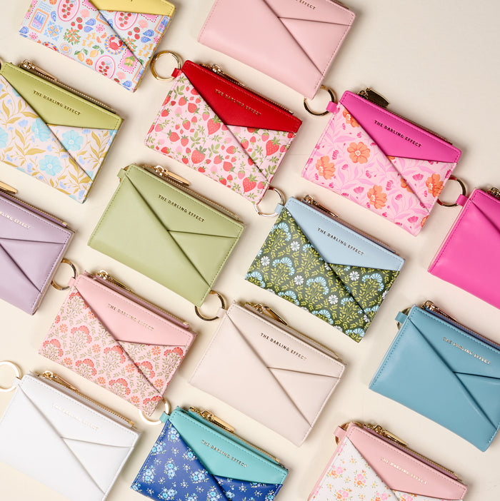 The Darling Effectβs White Luxe Pocket Wallet, crafted from vegan leather, features a gold zipper and key ring. Shown with other compact wallets and cardholders in pastel and floral patterns, neatly arranged on a light background.