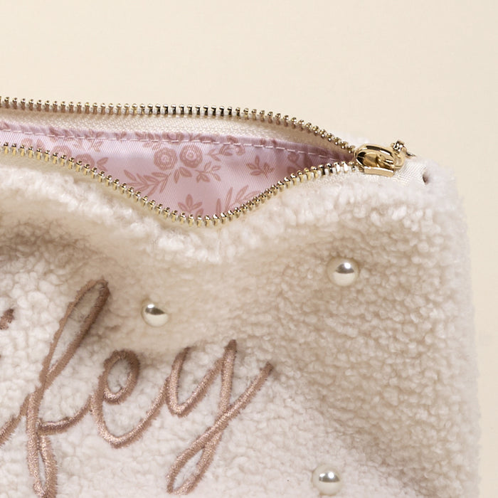 The Darling Effectโs Wifey & Pearls Teddy Pouch features soft cream sherpa, decorative pearls, light brown โWifeyโ embroidery, a gold zipper, and a pink floral inner lining.