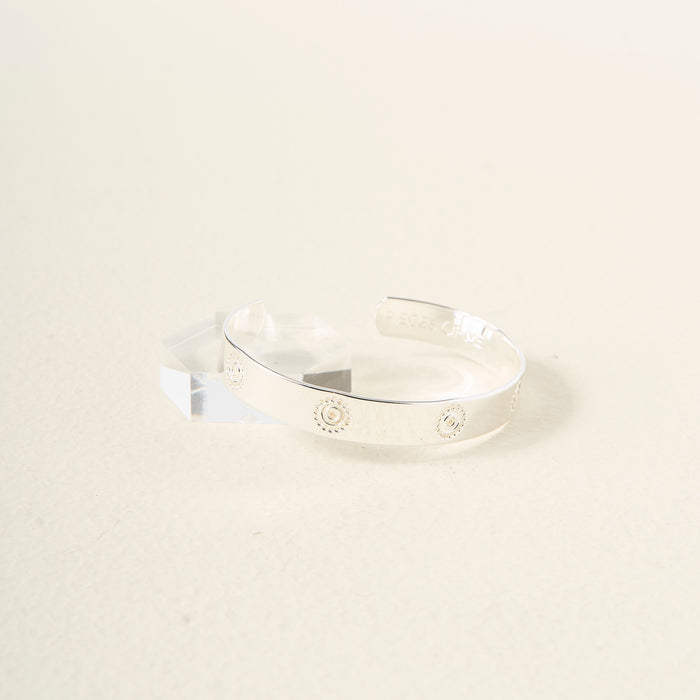 The Darling Effect's You Are Confident Trait Cuff Bracelet, crafted in sterling silver with engraved sunburst designs, rests on a light textured surface beside a clear geometric blockβmaking it a meaningful gift for any occasion.