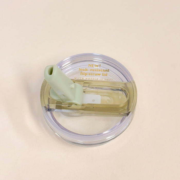 A top view of The Darling Effect 20 oz Green Lifestyle Flip Straw Replacement Lid features a clear plastic lid with a light green silicone straw, shown on a light beige background. Gold text reads: NEW! leak-resistant seal flip straw lid—perfect for replacements.