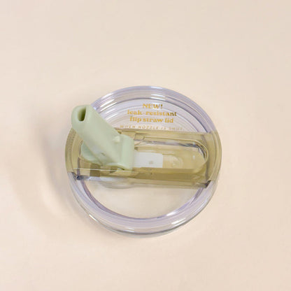 A top view of The Darling Effect 20 oz Green Lifestyle Flip Straw Replacement Lid features a clear plastic lid with a light green silicone straw, shown on a light beige background. Gold text reads: NEW! leak-resistant seal flip straw lid—perfect for replacements.