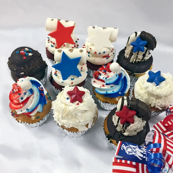Nine 4th of July–themed assorted cupcakes arranged on a light surface, each topped with red, white, and blue frosting, sprinkles, star-shaped toppers or scroll decorations.