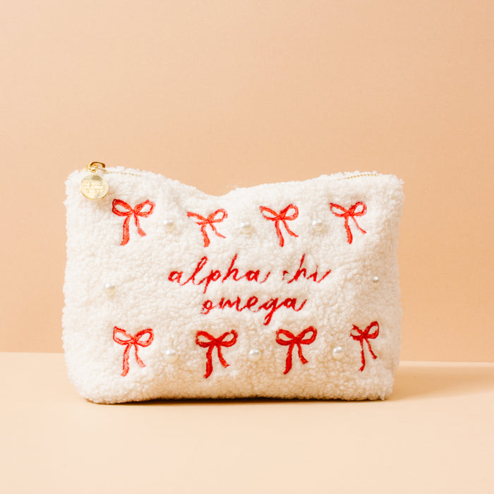 The Darling Effect’s Alpha Chi Omega Bows & Pearls Teddy Pouch features a white sherpa texture, red embroidered bows, pearl details, and “alpha chi omega” stitched in red—an ideal sorority gift against a peach background.