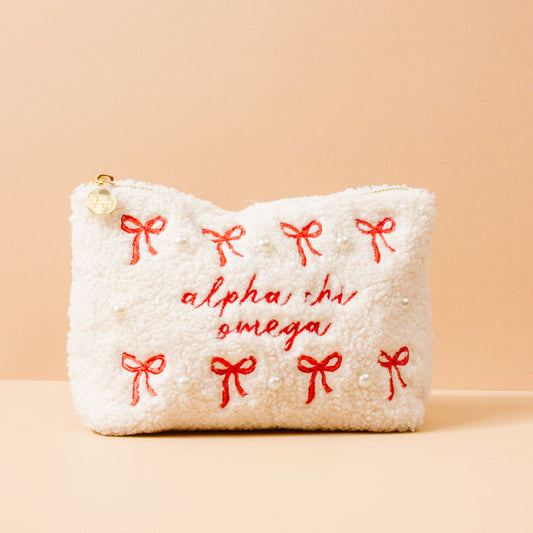 The Darling Effect’s Alpha Chi Omega Bows & Pearls Teddy Pouch features a white sherpa texture, red embroidered bows, pearl details, and “alpha chi omega” stitched in red—an ideal sorority gift against a peach background.
