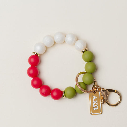 The Darling Effect’s Alpha Chi Omega Hands-Free Keychain Wristlet features red, green, and white round beads in a circle, a gold keyring and clasp, plus a rectangular Greek letter charm engraved with ΑΧΩ.