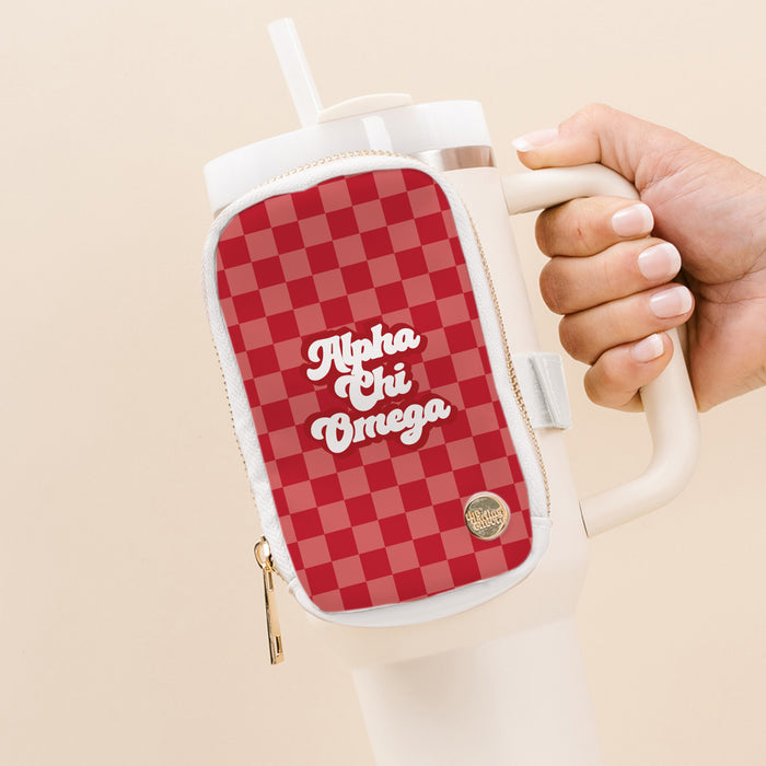 A hand holds a white tumbler with a red checkered, water-resistant Alpha Chi Omega Tumbler Fanny Pack by The Darling Effect, featuring “Alpha Chi Omega” in white script, a zipper pouch, and a small gold emblem near the bottom.