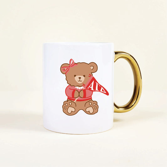 The Darling Effect's Alpha Gamma Delta 11 oz Cozy Cute Ceramic Mug features a ceramic build and gold handle, adorned with a teddy bear in a red bow, necklace, sweater, and holding an Alpha Gamma Delta pennant—perfect for proud members!.