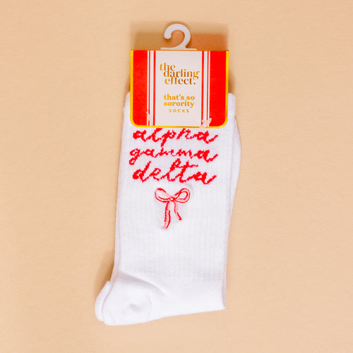 Show your sorority spirit with The Darling Effect Alpha Gamma Delta Embroidered Crew Socks—white crew socks featuring red script and a red bow, packaged on a peach background.